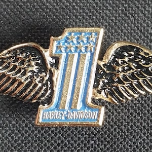 May include: A gold-colored metal pin featuring a stylized number one with blue and white stripes and stars, flanked by black and gold wings. The pin has the text "HARLEY-DAVIDSON" at the base.