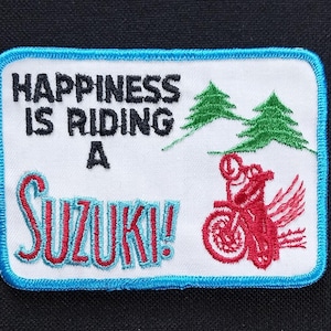 Puede incluir: Un parche bordado azul y blanco con el texto "Happiness is riding a Suzuki!" y una motocicleta roja con un conductor. El parche presenta árboles verdes en el fondo.