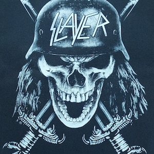 Op de afbeelding: Zwart grafisch ontwerp met een schedel met een helm met het woord "SLAYER" in wit. Twee gekruiste zwaarden staan achter de schedel. De schedel heeft lang haar en een dreigende uitdrukking. Afbeelding voor bandmerchandise.