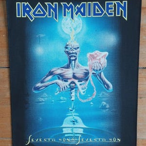 以下が含まれることがあります： 青いブロック体の文字で「IRON MAIDEN」と書かれた黒い長方形のパッチ。画像には、ピンク色の物体を持つ骸骨の姿が描かれており、その下に「SEVENTH SON OF A SEVENTH SON」という文字があります。デザインは青と白の背景です。