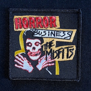 Pode incluir: Um patch quadrado preto com as palavras "HORROR BUSINESS" e "THE MISFITS". O patch apresenta um gráfico de caveira com detalhes vermelhos e brancos, e detalhes amarelos e brancos. O design é bordado.