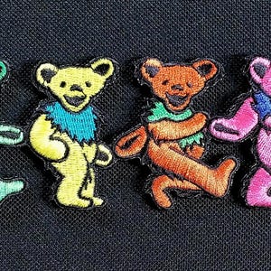 May include: A row of six embroidered dancing bear patches in various colors: blue, green, yellow, orange, pink, and purple. Each bear has a different colored collar and is walking with its arms and legs outstretched.