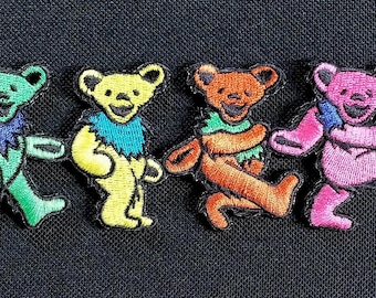 May include: A row of six embroidered dancing bear patches in various colors: blue, green, yellow, orange, pink, and purple. Each bear has a different colored collar and is walking with its arms and legs outstretched.