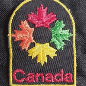 May include: A black embroidered patch with a yellow border featuring four maple leaves in red, orange, green, and yellow arranged in a circular pattern around a center hole. The word Canada is stitched in red letters at the bottom.