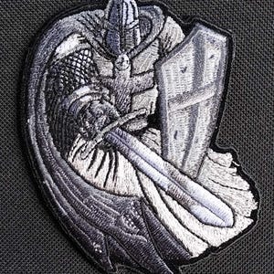 May include: A black and white embroidered patch of a knight in armor holding a sword and shield. The knight is wearing a helmet and a cape. The shield has a cross on it.