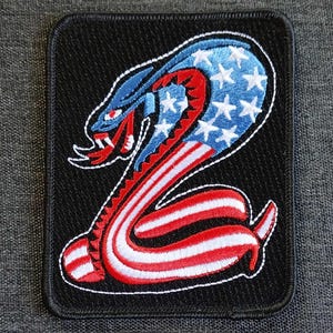 May include: A black embroidered patch with a red, white, and blue cobra design. The cobra is coiled and has a fierce expression. The cobra's scales are patterned with the American flag.