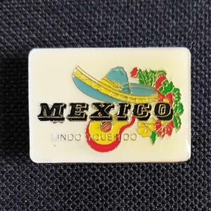 May include: A white rectangular pin featuring a colourful image of a sombrero, guitar, and flowers. The text "MEXICO" is in black lettering and "LINDO QUERIDO" is in smaller black lettering.