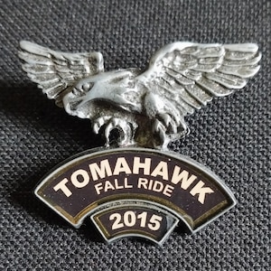 May include: A silver eagle pin with a black and gold banner that reads "Tomahawk Fall Ride 2015".
