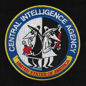 Puede incluir: Parche bordado circular con borde azul y las palabras "CENTRAL INTELLIGENCE AGENCY" y "UNITED STATES OF AMERICA". El parche presenta dos espías de dibujos animados, uno negro y otro blanco, sosteniendo una bomba y un cartucho de dinamita.