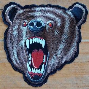 Peut inclure: Un patch brodé représentant un ours brun avec une expression féroce. La gueule de l'ours est ouverte, révélant des dents acérées et une langue rouge. Les yeux de l'ours sont rouges et intenses.