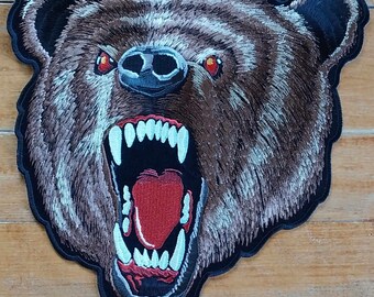 Puede incluir: Un parche bordado que representa un oso marrón con una expresión feroz. La boca del oso está abierta, mostrando dientes afilados y una lengua roja. Los ojos del oso son rojos e intensos.