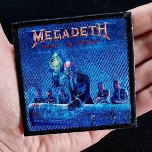 Può includere: Una toppa ricamata blu e nera con il nome della band "Megadeth" e il titolo dell'album "Rust in Peace". La toppa presenta un membro della band che tiene una lampadina e altri membri della band sullo sfondo.