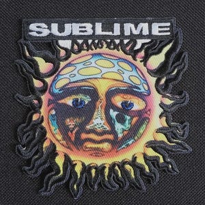 Op de afbeelding: Geborduurde patch met een gestileerde zon met een gezicht, met een blauw en geel patroon hoofdbedekking. De zon heeft blauwe ogen en een kleurrijk ontwerp. Het woord "SUBLIME" is in witte letters boven de zon geborduurd.
