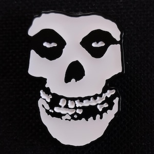 以下が含まれることがあります： 白と黒のエナメルピン。ミズフィッツの象徴的なスカルロゴが描かれています。スカルは黒い眼窩、黒い鼻、そして大きく歯を見せた笑顔をしています。ピンは黒い背景に配置されています。