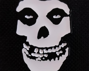 Puede incluir: Un pin de esmalte blanco y negro con el icónico logotipo de la calavera de Misfits. La calavera tiene cuencas de ojos negras, una nariz negra y una amplia sonrisa con dientes. El pin está sobre un fondo negro.