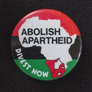 May include: A round button with a red, black, and green design. The button features a white outline of the African continent with the words "ABOLISH APARTHEID" and "DIVEST NOW" in white text.