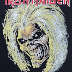 May include: Black graphic featuring the Iron Maiden band logo in red and a detailed illustration of the band's mascot, Eddie. Eddie is depicted as a skeletal figure with yellow hair and a menacing expression.