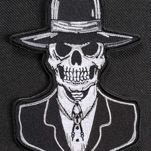 May include: A black and white embroidered patch of a skull wearing a fedora and a suit. The skull is wearing a tie with a bolo tie clasp.