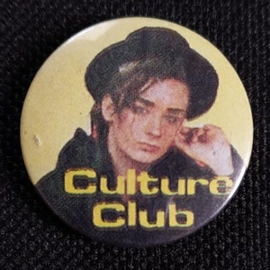 May include: A yellow and black button featuring a photo of a man wearing a black hat and a black jacket. The text "Culture Club" is printed in yellow letters on the button.