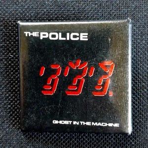 May include: Square black button featuring the band name "THE POLICE" in white text above a red digital display of the number "333". Below the number is the text "GHOST IN THE MACHINE".
