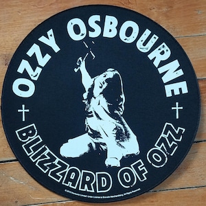 Puede incluir: Un parche redondo negro con las palabras "OZZY OSBOURNE" y "BLIZZARD OF OZZ" en blanco. Presenta una ilustración blanca de una persona sosteniendo una cruz. Hay cruces a ambos lados del nombre.