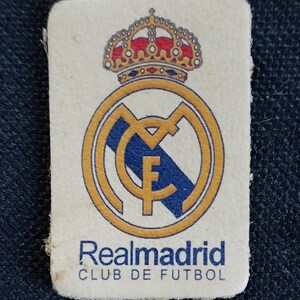 May include: A white patch with a gold and blue Real Madrid logo. The logo features a crown, a circle with a letter M, and a blue and white stripe. The text "Realmadrid CLUB DE FUTBOL" is printed below the logo.