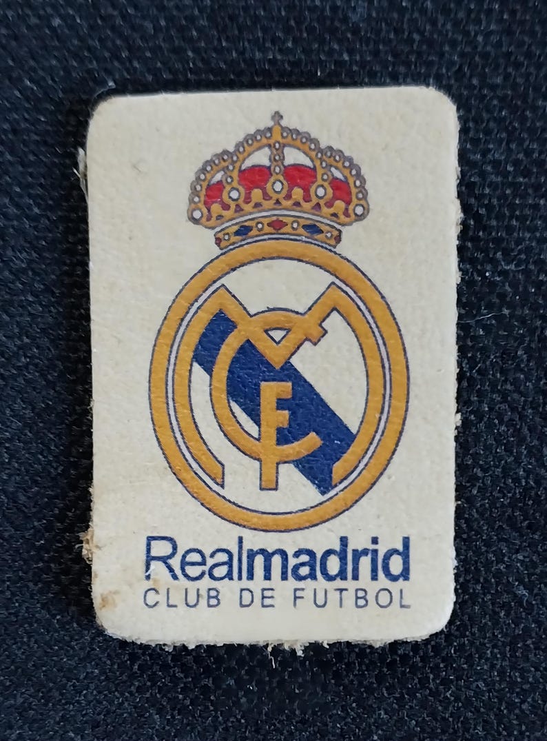 May include: A white patch with a gold and blue Real Madrid logo. The logo features a crown, a circle with a letter M, and a blue and white stripe. The text "Realmadrid CLUB DE FUTBOL" is printed below the logo.