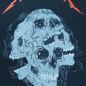 Könnte beinhalten: Schwarzes T-Shirt mit einem hellblauen Schädeldesign mit mehreren sich überlappenden Schädeln. Das Wort "METALLICA" ist oben in Orange aufgedruckt. Das Design ist eine grafische Illustration. Copyright-Informationen unten.