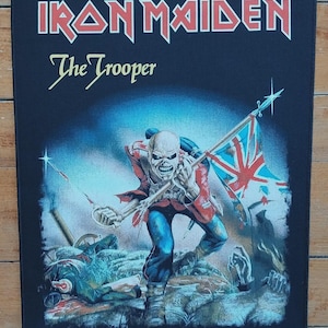 Puede incluir: Parche rectangular negro con el texto "IRON MAIDEN" en rojo y blanco, y "The Trooper" en dorado. El gráfico muestra una figura esquelética con abrigo rojo y pantalones azules, sosteniendo una bandera de la Union Jack, en un campo de batalla.