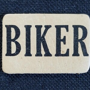 May include: A beige leather patch with the word "BIKER" printed in black.