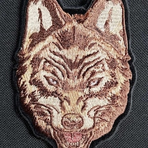 May include: Embroidered patch of a wolf's head in brown and beige tones. The wolf's mouth is open, showing its teeth.