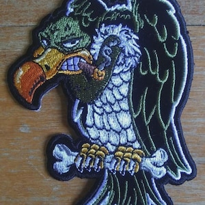 May include: Embroidered patch featuring a cartoon vulture with a menacing expression. The vulture has a green head, orange beak, and is clutching a bone. The patch is outlined in black with green, white, and yellow accents.