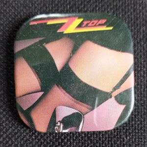 May include: A square button with a black and white image of legs in stockings and a high heel shoe. The top of the button has the text "ZZ Top" in yellow and red letters.