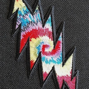 Puede incluir: Un parche bordado con forma de rayo que presenta un vibrante patrón tie-dye con remolinos de hilo rojo, amarillo, azul y blanco. El rayo tiene un borde grueso cosido en negro y se encuentra sobre un fondo de tela negra texturizada.