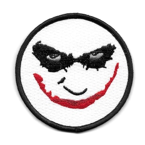 Peut inclure: Un &eacute;cusson brod&eacute; blanc avec un contour noir, repr&eacute;sentant un visage de type Joker avec un sourire rouge et des yeux noirs.