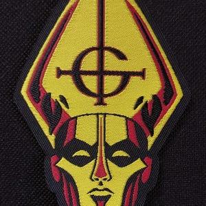 May include: A yellow, red, and black patch featuring a stylized face and a symbol. The patch has a diamond shape with a yellow background and a red and black outline. The face has a red outline and a yellow center. The symbol is black.