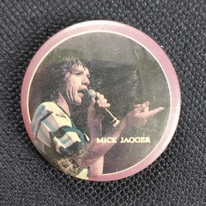 Könnte beinhalten: Ein runder Knopf mit rosa Rand und einem Schwarzweißbild eines Mannes, der in ein Mikrofon singt. Der Text "MICK JAGGER" ist in Weiß unter dem Bild gedruckt.