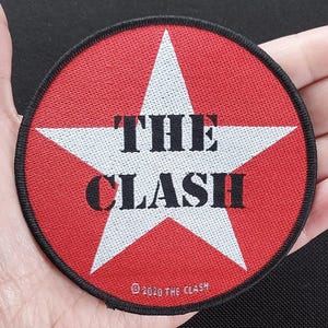 Peut inclure: Un patch circulaire rouge et blanc avec une étoile blanche et le texte "THE CLASH" en lettres noires. Le texte "©2010 THE CLASH" est imprimé en noir au bas du patch.