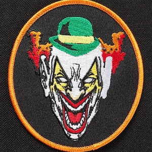 May include: Circular embroidered patch depicting a clown's face with a wide, red mouth. The design features a green hat, yellow details, and orange edging on a black backdrop.