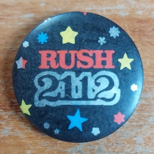 May include: A black button featuring the word "RUSH" in red and orange lettering, with the year "2012" in silver lettering. The button is adorned with colourful stars.