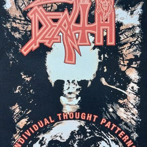 May include: Black t-shirt with the band name "DEATH" in a red, stylized font. The design includes a skull and the words "INDIVIDUAL THOUGHT PATTERNS" in red. The background has a dark, textured appearance.
