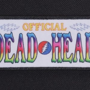 May include: A rectangular patch with the words "OFFICIAL DEAD HEAD" in colorful, psychedelic lettering. The patch features a Steal Your Face logo and is bordered by a black frame.