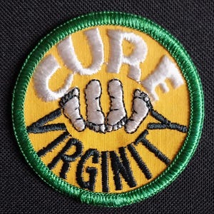 May include: A yellow and green embroidered patch with the words "Cure Virginity" in white and black lettering. The patch features a stylized image of three pairs of feet.