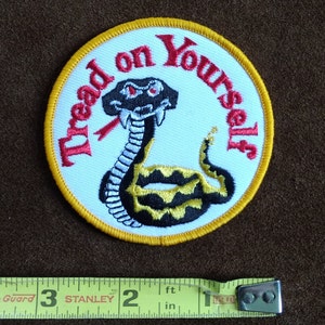 Può includere: Una toppa ricamata bianca con un bordo giallo. La toppa presenta un serpente a sonagli nero e giallo con occhi rossi e il testo "Tread on Yourself" in rosso.