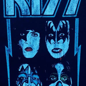 May include: Black t-shirt featuring the band KISS. The design includes the band's logo in light blue at the top, with four band member faces below. Each face has unique makeup. Lightning bolts frame the design.