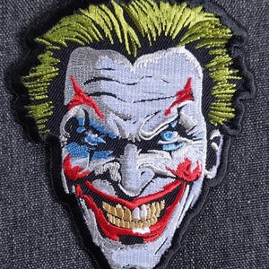 May include: Embroidered patch of a white-faced, grinning joker with red lips, blue eyes, and yellow teeth. The joker has a green and yellow mohawk hairstyle.