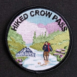May include: A round, black and white embroidered patch with the text "HIKED CROW PASS" in a circle around a scene of a hiker walking along a stream in a mountain valley. The stream has a sign that reads "WAN PATCH".