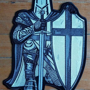 Puede incluir: Un parche bordado en blanco y negro de un caballero arrodillado con una espada y un escudo. El escudo tiene una cruz blanca sobre un fondo negro.