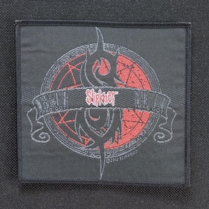 Puede incluir: Parche cuadrado negro con el nombre de la banda "Slipknot" en rojo, con un emblema y una pancarta estilizados. El diseño incluye un pentagrama rojo y una fuente gótica. El parche tiene un borde negro.