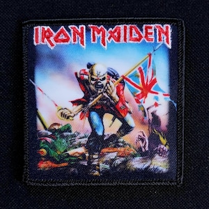 Könnte beinhalten: Ein quadratischer Aufnäher mit dem Bandnamen "Iron Maiden" in roten Buchstaben. Das Bild zeigt eine skelettartige Figur in einem roten Mantel und blauen Hosen, die ein Schwert und eine Union Jack-Flagge hält. Der Hintergrund zeigt eine Schlachtfeldszene.